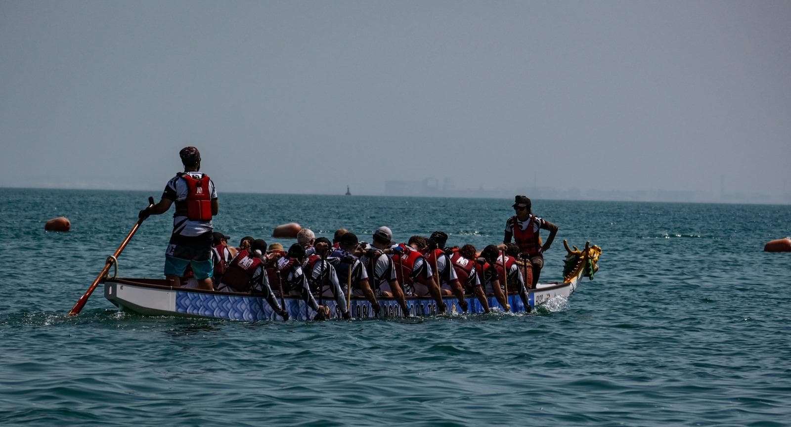 A dragon boat crew paddling on open water, with the steersperson standing at the stern and the drummer at the bow